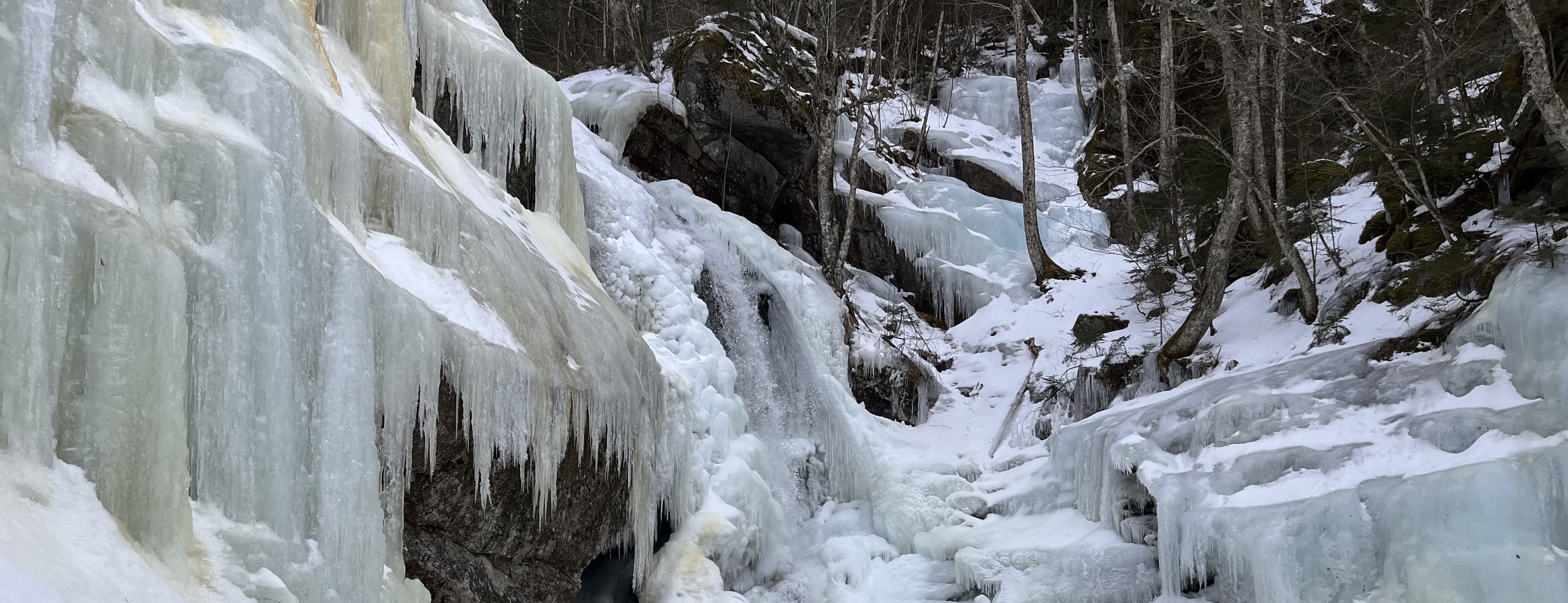 A frozen waterfall in the northern New Hampshire woods, ice stacked in sheets over the rock